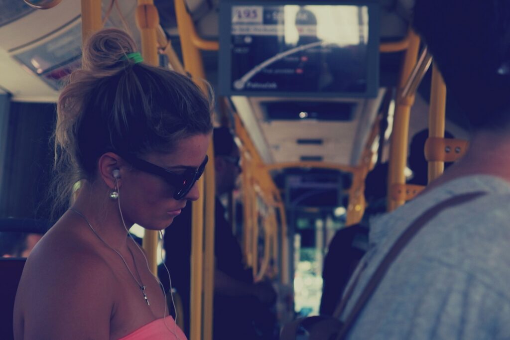 A woman with her hair in a bun wears sunglasses and ear buds on a public bus.